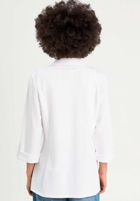 Blazer blanc à manches trois-quarts, col et revers retroussés. Tissu lisse, coupe légèrement cintrée, et fentes latérales pour faciliter les mouvements.