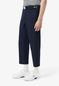 Pantalons bleu marine à coupe droite, avec une ceinture noire à boucle argentée et un logo vert. Associés à des chaussures de sport blanches.
