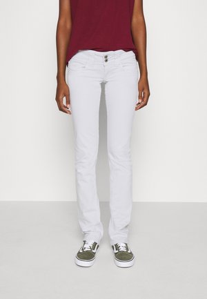 Persona con pantalones blancos ajustados, top rojo oscuro y zapatillas verdes y blancas, de pie frente a un fondo claro y sencillo.