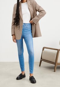 Persona vestida con jeans ajustados de color azul claro, mocasines negros, camisa blanca y blazer beige, de pie cerca de una silla de madera con cojín beige.