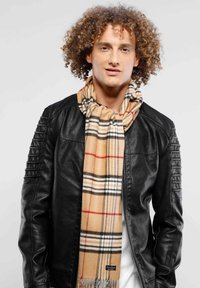 Beige plaid scarf with red and black accents, fringed ends, draped around neck, worn over a black leather jacket with textured sleeves.