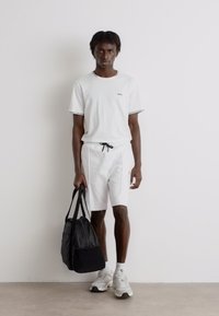 T-shirt blanc à manches courtes et shorts avec bord noir, associés à un sac de sport noir et des chaussures de sport grises. Tissu lisse, design décontracté.