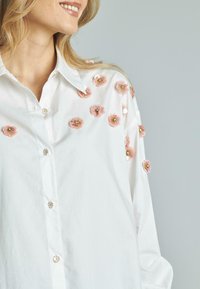 Camicia bianca con applicazioni floreali rosa e decorazioni, con bottoni ornati da piccoli motivi di margherite sulla parte anteriore.