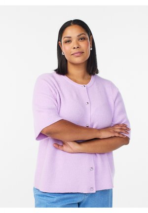 Kvinde med glat mørkt hår, iført sølvfarvede øreringe i ringform, en lilla knappet kortærmet sweater og lyseblå jeans, med armene krydset.