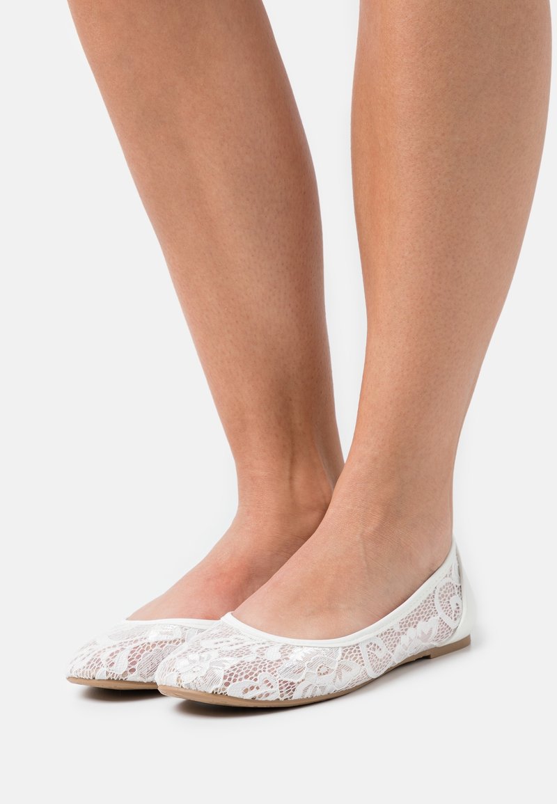 Jambes portant des ballerines en dentelle blanche avec des motifs floraux et des semelles marron clair, présentées sur un fond blanc uni.