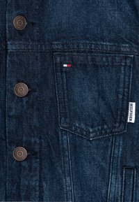 Giubbotto in denim scuro con bottoni in rame, una tasca sul petto con un piccolo etichetta rossa, bianca e blu, e una finitura leggermente testurizzata.