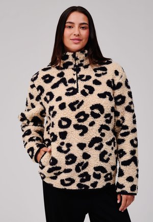 Femme portant un pull polaire beige et noir à motif léopard avec une fermeture éclair à l'avant et les mains dans les poches, debout devant un fond uni.