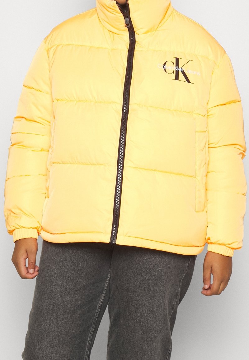 Giacca a vento gialla con chiusura a zip, colletto alto e polsini elasticizzati. Presenta il logo Calvin Klein sul lato sinistro del petto e un design trapuntato.