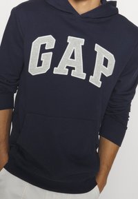 Person som bär en marinblå huvtröja med stora grå "GAP"-bokstäver över bröstet och händerna delvis i fickorna på ljusa byxor.