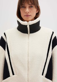 Weiße Fleecejacke mit schwarzen Akzenten und hohem Kragen. Sie verfügt über einen Frontreißverschluss und geometrische Muster in plüschiger Textur.