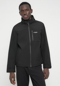 Black softshell jacket with high collar, front zipper, and chest pocket. Features a minimalist design with subtle logo accents.