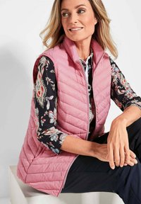 Helles rosa gesteppter Weste mit Stehkragen. Trägt sie über einem schwarzen Hemd mit Floral-Muster. Modell sitzt mit Händen auf den Knien.
