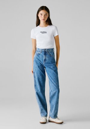 Giovane donna in piedi, che indossa una maglietta bianca "Red Bull Racing", jeans blu a vita alta e sneakers bianche, su uno sfondo grigio neutro.