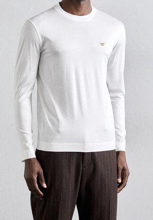 Homme portant une chemise blanche cintrée à manches longues avec un petit logo doré sur la poitrine et un pantalon à fines rayures brun foncé.
