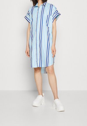 Frau trägt ein lockeres, kurzärmeliges Kleid in Knielänge mit blauen vertikalen Streifen und weißen Plateausneakern, steht vor einem schlichten Hintergrund.