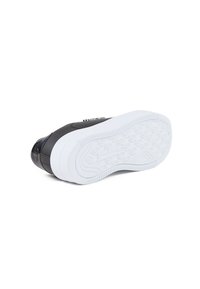 Zapatilla negra con parte superior suave, puntera texturizada y suela de goma blanca con patrones de agarre circulares. Marca sutil en el lateral.