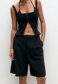 Kobieta w czarnym topie bez rękawów z zapięciem z przodu oraz czarnych, szerokich szortach do kolan, stojąca na białym tle.