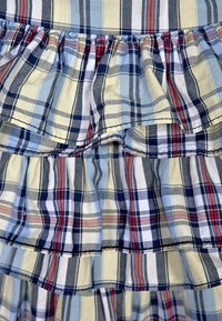 Károvaná tkanina s měnícími se modrými, žlutými a červenými vzory. Design zahrnuje vrstvené volány se viditelnými švy.