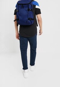 Osoba w czarno-niebieskiej koszuli, ciemnoniebieskich spodniach, białych sneakersach, niosąca dużą granatową plecak z bocznymi paskami, odchodzi na białym tle.