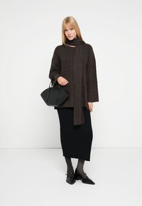Woman wearing a brown cable-knit sweater and scarf, black ribbed midi skirt, black tights, black shoes, and holding a black handbag.