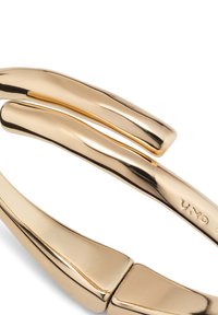 Pulsera de metal tono dorado con un diseño curvado y suave; presenta un distintivo doble lazo y una marca sutil.