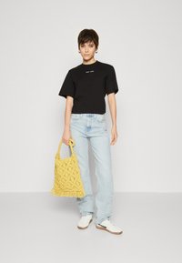 Černý krátký rukávový crop top s textovým logem; světle modré rovné džíny; hořčičně žlutá háčkovaná taška s třásněmi; bílé tenisky.