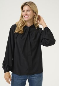 Blusa nera con colletto alto e dettaglio a fiocco, caratterizzata da maniche lunghe a sbuffo con polsini elastici e una vestibilità morbida. Tessuto dalla superficie liscia.