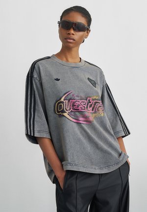 Άτομο που φοράει υπερμεγέθη γκρι μπλουζά Adidas με πολύχρωμο κείμενο "Questra", μαύρο αθλητικό παντελόνι και σκούρα γυαλιά ηλίου που τυλίγονται γύρω, με τα χέρια στις τσέπες.