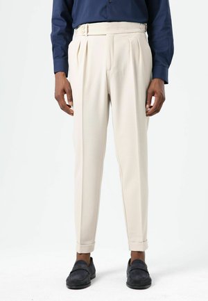 Pantalon beige plissé en tissu texturé, avec deux boutons à la taille et ourlets retroussés, porté avec des mocassins foncés.