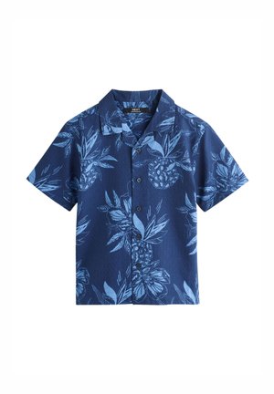 Garçon aux cheveux bouclés portant une chemise bleue à motifs floraux et un short blanc, assis à l'extérieur sous un ciel partiellement nuageux.