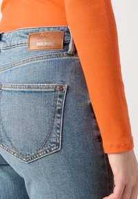 Jeans in denim blu con una texture liscia, caratterizzati da una toppa in pelle marrone con l'etichetta "MOS MOSH" e cuciture classiche, abbinati a un top arancione.
