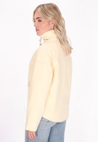 Jauna moteris su šviesiais plaukais, dėvinti šviesiai geltoną striukę, pasukusi dešinėn prieš šviesų foną, demonstruojanti minimalistinį stilių.