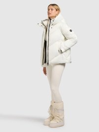 Veste blanche matelassée avec un col montant, fermeture éclair, et des accents noirs. Associée à des leggings crème et des bottes d'hiver beige montant jusqu'au genou.