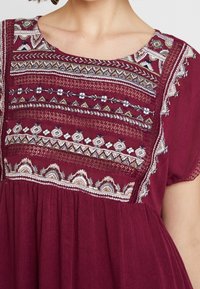 Burgundské šaty s jemnou výšivkou květinových a geometrických vzorů přes živůtek; lehká tkanina; krátké rukávy.