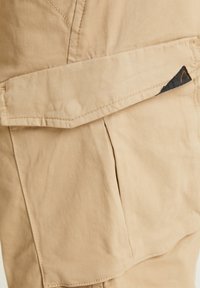 Tissu beige avec une poche rabat et un accent triangulaire noir. Texture lisse avec des détails de couture visibles.