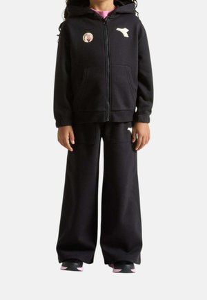 Bambino con giacca nera con cappuccio e zip e pantaloni a gamba larga con loghi bianchi e scarpe da ginnastica rosa su sfondo bianco.