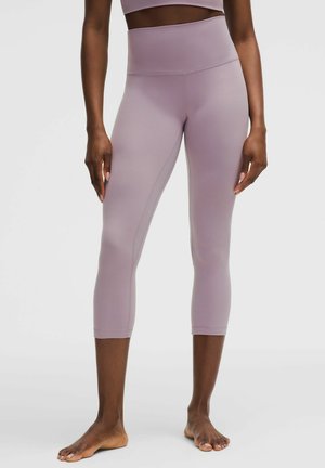 Persoon die mouwloze, hooggetailleerde, mauve-kleurige cropped leggings draagt, staat blootsvoets tegen een effen achtergrond.
