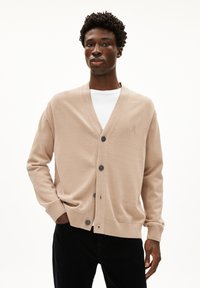 Beige cardigan van zachte brei- stof, voorzien van een V-hals, zes knopen, ribgebreide manchetten en een subtiele logo-details op de borst. Draagt over een wit shirt.