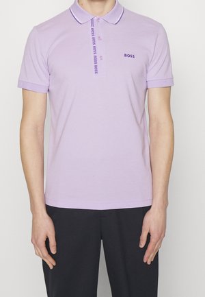 Mann trägt ein hellviolettes BOSS-Poloshirt mit Knopfleiste und gesticktem Logo, kombiniert mit schwarzen Hosen, steht vor einem weißen Hintergrund.
