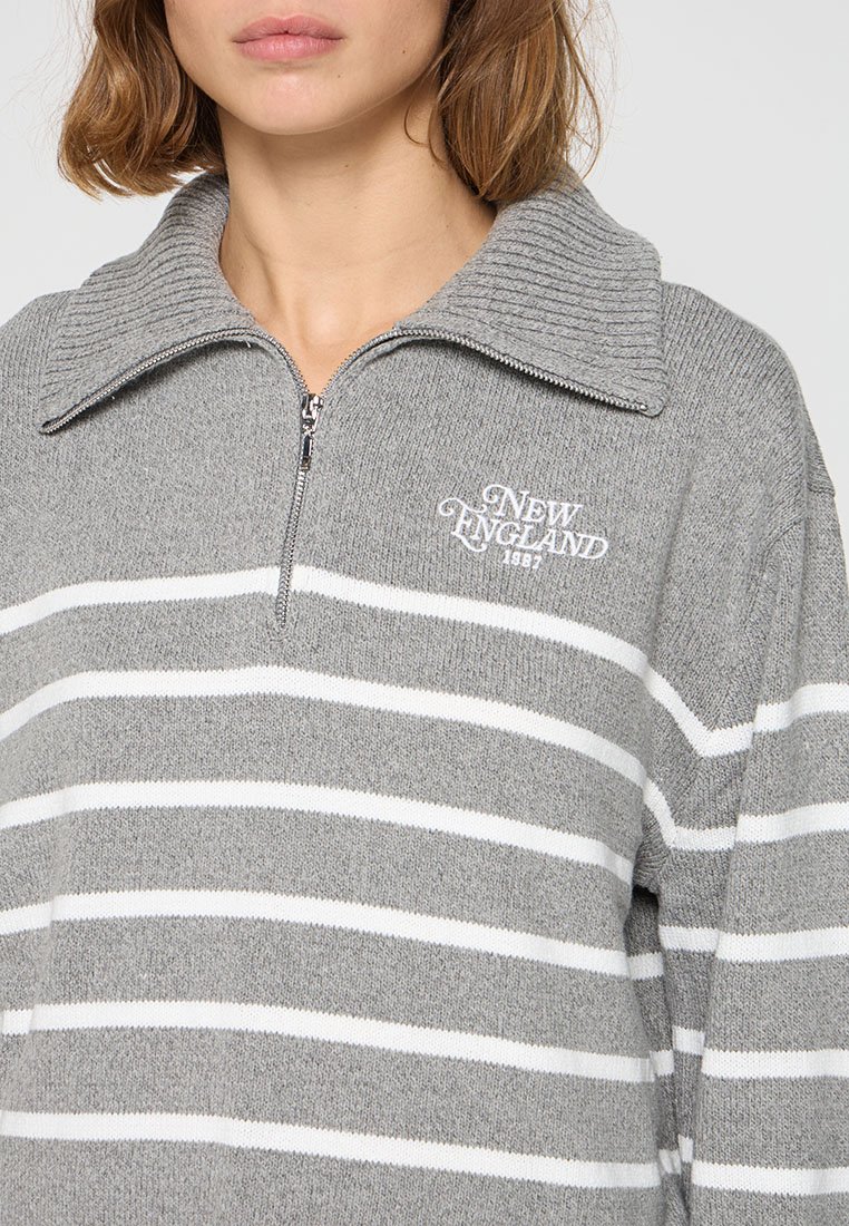 Sweatshirt zippé gris avec un col côtelé, présentant des rayures horizontales blanches et un logo "New England 1987" en broderie contrastante.