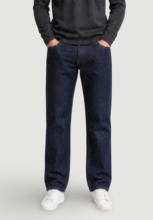 Hombre con vaqueros azul oscuro de pierna recta, camiseta negra de manga larga, zapatillas blancas, con las manos en los bolsillos de los vaqueros, de pie sobre fondo liso.