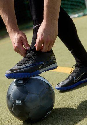 Scarpe da calcio nere con accenti blu, sopra un pallone da calcio bianco e nero, collocate su un campo verde. La tomaia è testurizzata e i tacchetti sono sagomati.