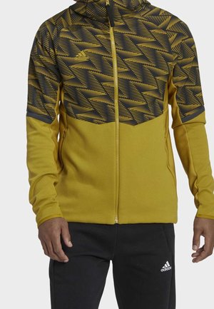 Homme portant une veste zippée à motifs jaune moutarde et noir et un pantalon Adidas noir avec le logo sur la cuisse, se tenant devant un fond uni.