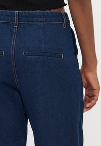 Dunkelblaue Jeans mit einer taillierten Passform, die horizontale Gesäßtaschen und Kontrastnähte aufweisen. Glatte Textur, hoher Taillenbund sichtbar.