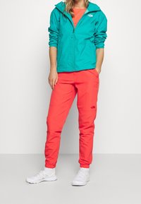 Nainen, joka pukeutuu meripihkansiniseen vetoketjulliseen takkiin, korallinvärisiin housuihin ja valkoisiin tennareihin, seisoo valkoista taustaa vasten käsi taskussa.