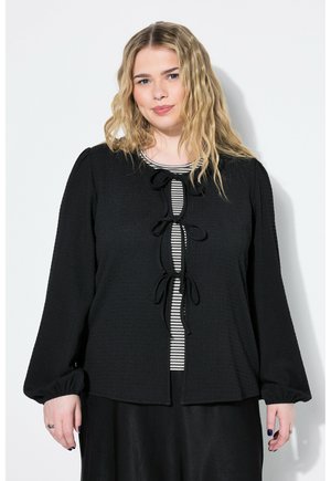 Sort tekstureret bluse med lange ærmer, med en nøglehulåbning prydet med lodrette sort og hvide striber, bundet med sløjfer.