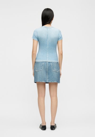 Γυναίκα που στέκεται με την πλάτη στην κάμερα, φορώντας ανοιχτό μπλε κοντομάνικο εφαρμοστό τοπ, τζιν φούστα με πίσω τσέπες και μαύρα παπούτσια με στρας.