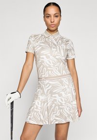 Golfové šaty s krátkým rukávem, béžová látka s bílým abstraktním vzorem, skládací sukně a límečkový výstřih, nošeny s golfovou rukavicí.