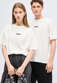 Νεαρή γυναίκα και άνδρας φοράνε λευκά μπλουζάκια με σχέδια, στέκονται δίπλα-δίπλα μπροστά σε απλό φόντο, η γυναίκα κρατάει ένα καρό πουκάμισο.