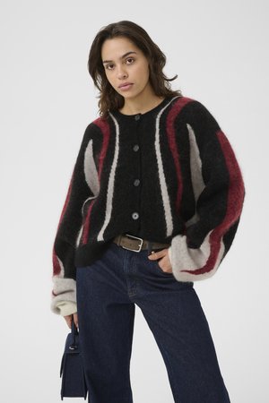 Woman wearing black, red, and white striped fuzzy cardigan, dark blue jeans, brown belt, holding small dark handbag, posing against light background.